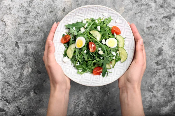 Vrouwelijke handen houden plaat met smakelijke rucola salade op grijze achtergrond — Stockfoto