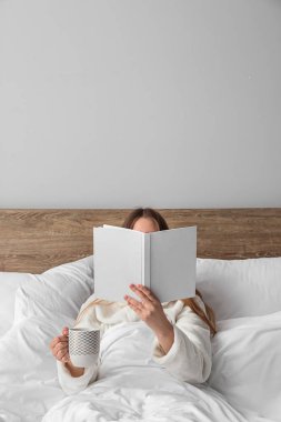 Genç bir kadın sıcak içecek bardağıyla yatakta kitap okuyor.