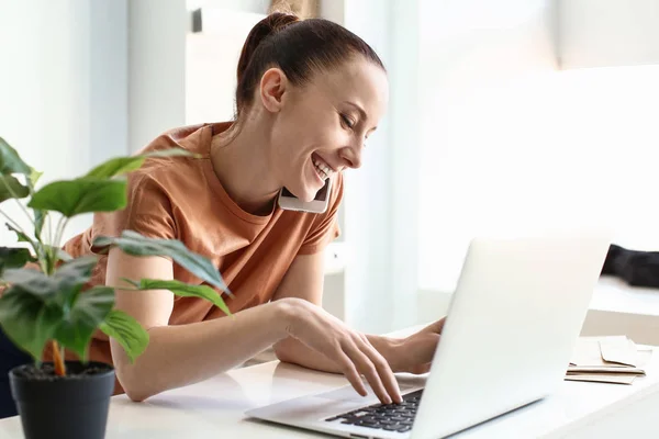 Woman talking by phone while working on laptop indoors — Stock Photo, Image