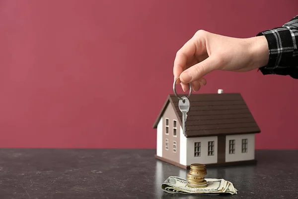 Frauenhand mit Schlüssel, Hausfigur und Geld auf dem Tisch vor farbigem Hintergrund. Konzept des Immobilienerwerbs — Stockfoto