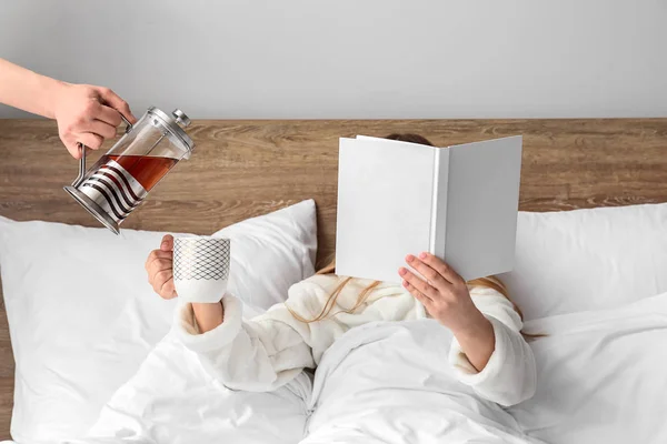 Maid pouring hot tea into cup of young woman sitting in bed — Stock Photo, Image