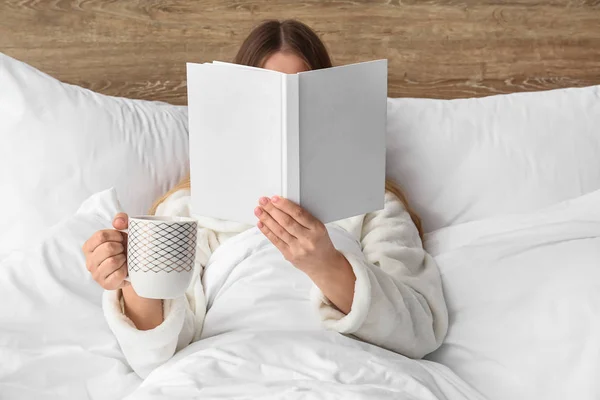 Young woman with cup of hot beverage reading book in bed — Stock Photo, Image