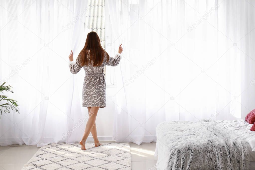 Young woman opening light curtains in bedroom at home