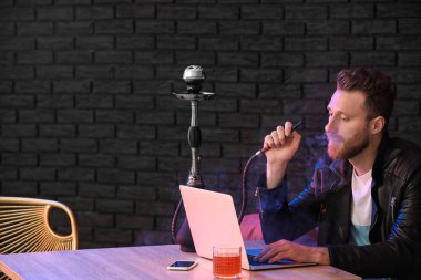 Genç adam kafede dizüstü bilgisayarla çalışırken nargile içiyor.