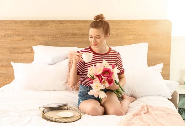 Mujer joven con ramo de hermosos tulipanes y taza de café sentado en la cama — Foto de Stock