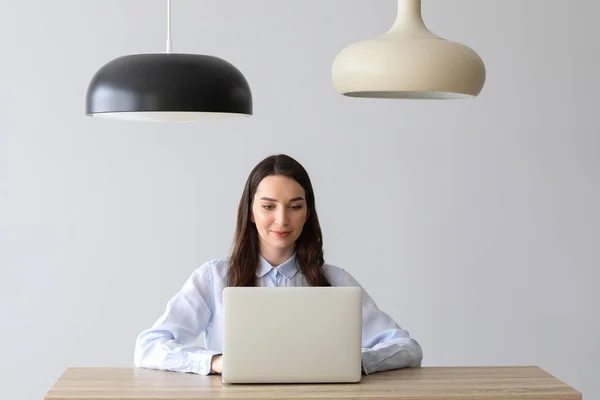 Vacker ung kvinna med bärbar dator sitter vid bordet mot ljus bakgrund — Stockfoto