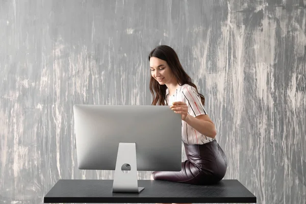 Beautiful young woman with computer sitting on table against grey background — Stock Photo, Image