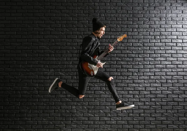 Salto giovane uomo con chitarra contro muro di mattoni scuri — Foto Stock