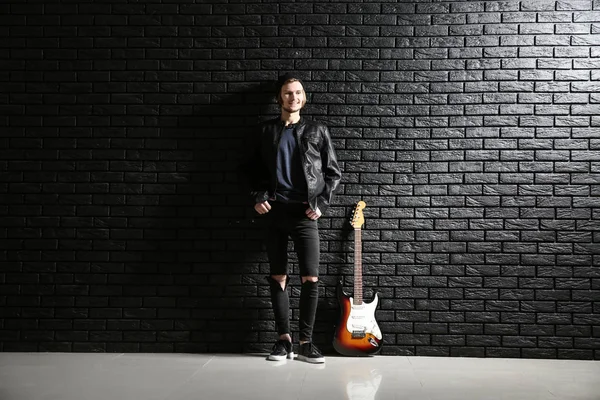 Bello giovane uomo con chitarra vicino muro di mattoni scuri — Foto Stock