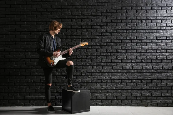 Bello giovane che suona la chitarra contro il muro di mattoni scuri — Foto Stock