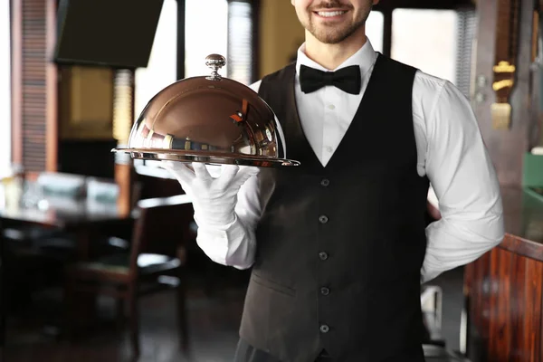 Young male waiter with tray and cloche in restaurant
