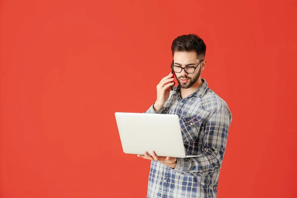 Male programmer with laptop talking by phone on color background — Stock Photo, Image