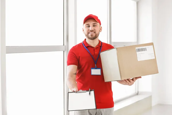 Handsome delivery man with box and clipboard indoors