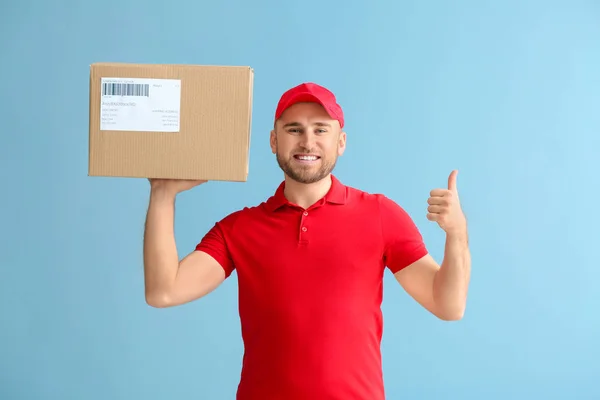 Handsome delivery man with box showing thumb-up on color background