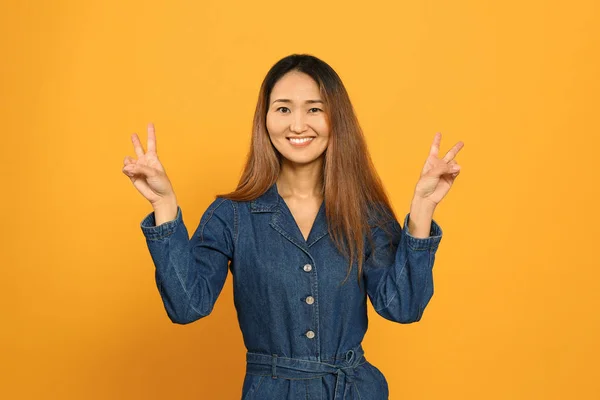 Portrait of Asian woman showing victory gesture on color background — Stock Photo, Image