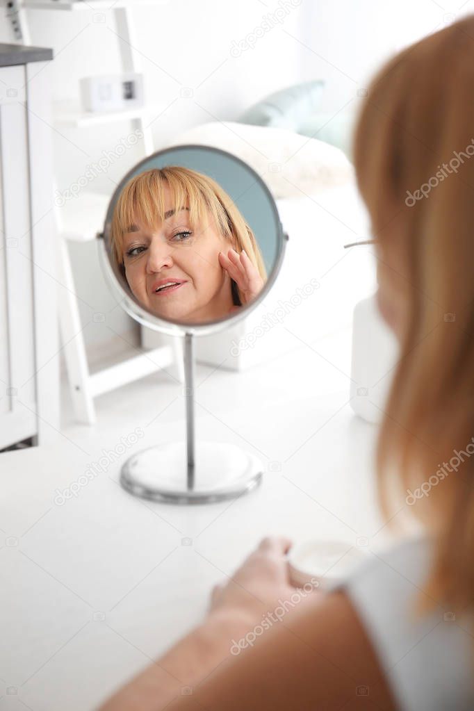 Mature woman with jar of cream looking in mirror at home