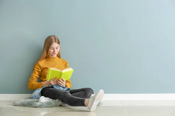 Симпатична дівчина-підліток з книгою, що сидить на підлозі біля кольорової стіни — стокове фото