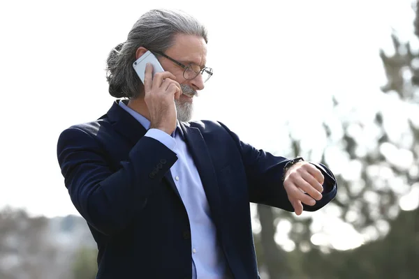 Knappe volwassen zakenman met mobiele telefoon kijken naar zijn horloge buitenshuis — Stockfoto