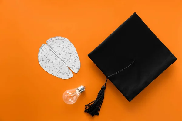 Mortar board with paper brain and light bulb on color background. Concept of high school graduation