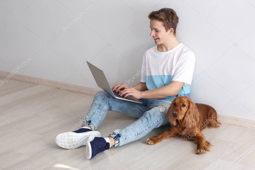 Teenage boy with cute dog and laptop sitting near light wall
