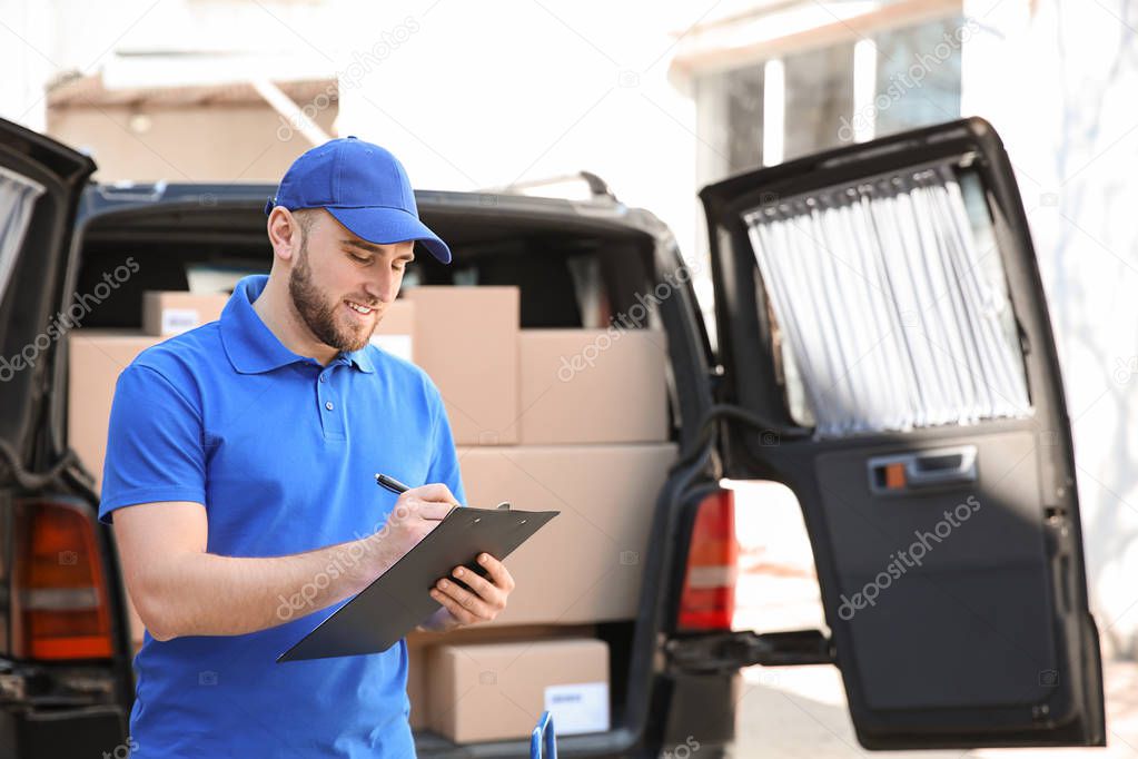 Handsome delivery man with clipboard near car outdoors