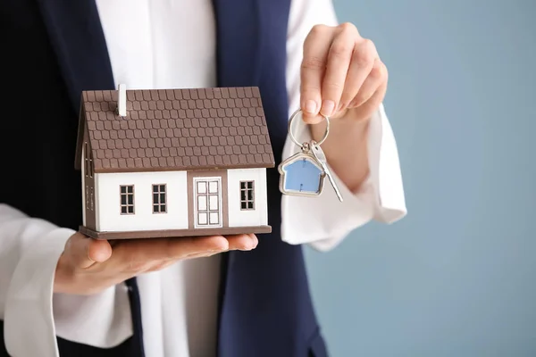 Female real estate agent with house model and key on grey background, closeup — Stock Photo, Image