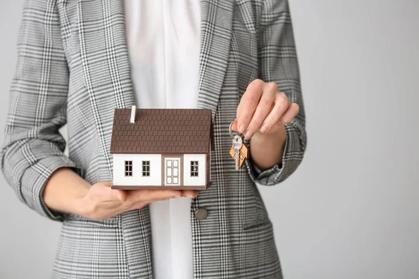 Female real estate agent with house model and key on grey background, closeup — Stock Photo, Image