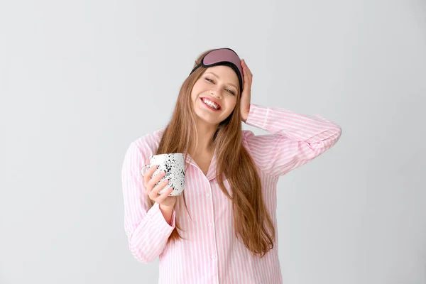 Beautiful young woman in pajamas and with cup of coffee on light background