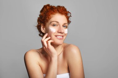 Beautiful redhead woman applying facial cream against grey background