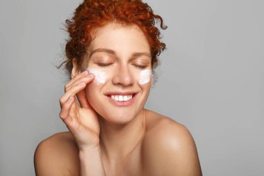 Beautiful redhead woman applying facial cream against grey background