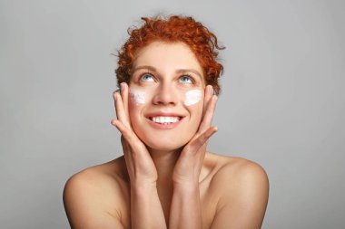 Beautiful redhead woman applying facial cream against grey background