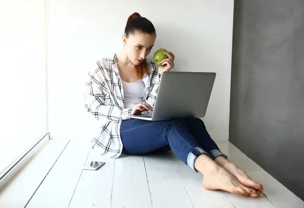 Молода жінка їсть яблуко під час роботи на ноутбуці в приміщенні — стокове фото