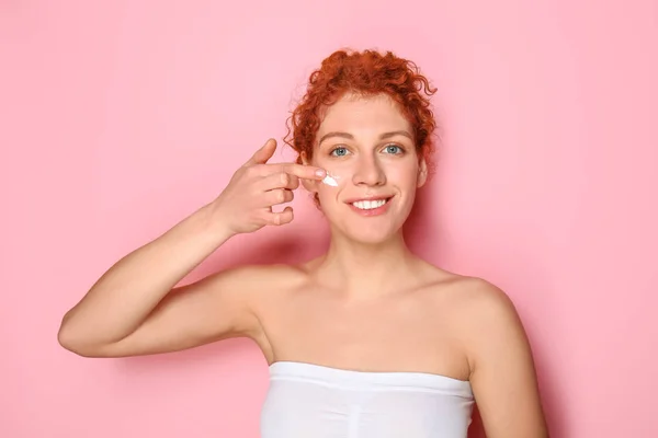 Beautiful redhead woman applying facial cream against color background — Stock Photo, Image