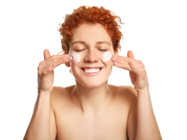 Hermosa pelirroja aplicando crema facial sobre fondo blanco — Foto de Stock