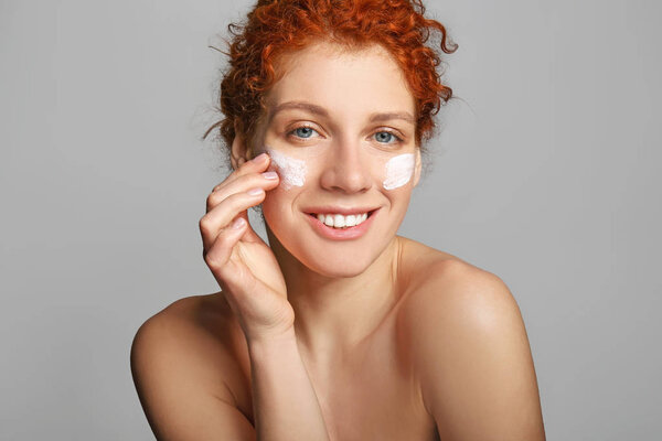 Beautiful redhead woman applying facial cream against grey background