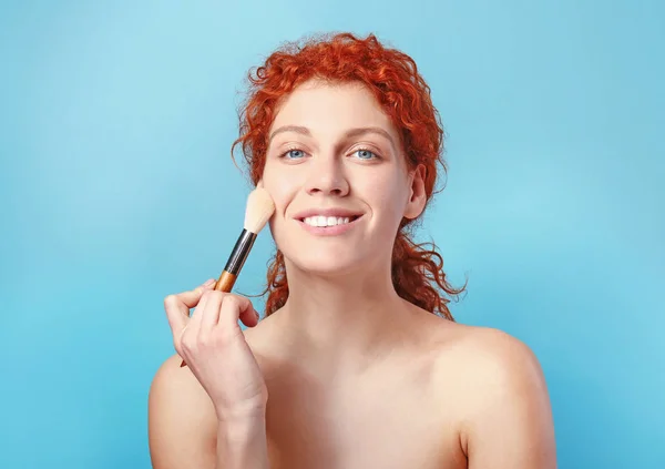 Beautiful redhead woman applying makeup against color background