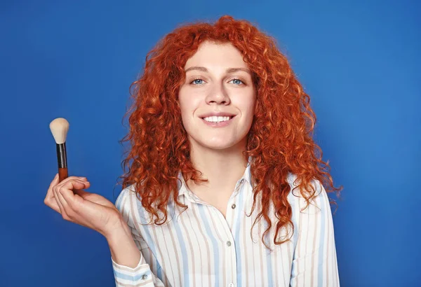 Beautiful redhead woman with makeup brush against color background