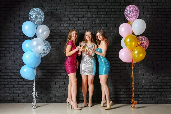 Beautiful young women with glasses of champagne and balloons near dark wall — Stock Photo, Image