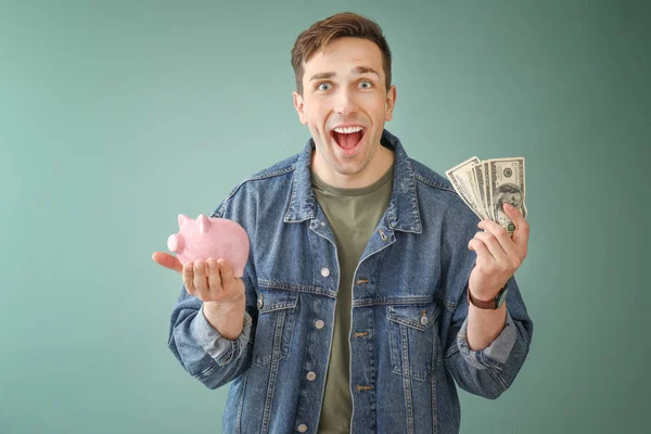 Happy young man with dollar banknotes and piggy bank on color background — Stock Photo, Image