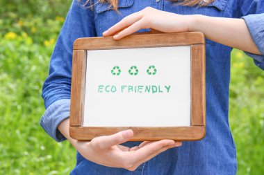Woman holding board with text ECO FRIENDLY outdoors, closeup
