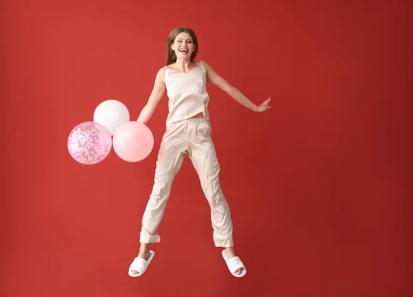 Jumping young woman in pajamas and with air balloons on color background — Stock Photo, Image