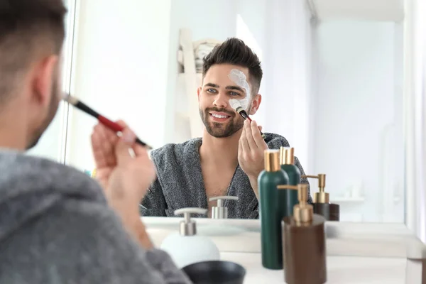 Hombre guapo aplicando máscara de arcilla en su cara en casa — Foto de Stock