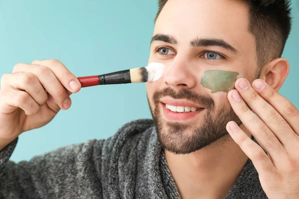 Schöner Mann, der vor farbigem Hintergrund Tonmaske auf sein Gesicht aufträgt — Stockfoto