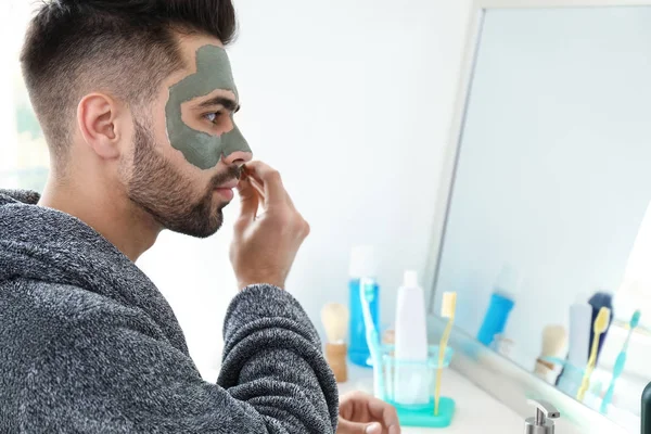 Bonito homem aplicando máscara de barro em seu rosto em casa — Fotografia de Stock