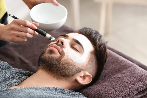 Cosmetologist applying clay mask onto face of young man in beauty salon — Stock Photo, Image