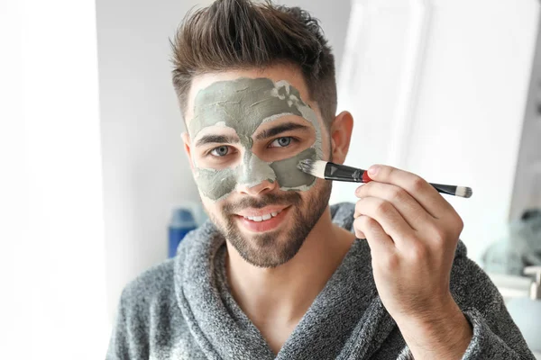 Hombre guapo aplicando máscara de arcilla en su cara en casa — Foto de Stock
