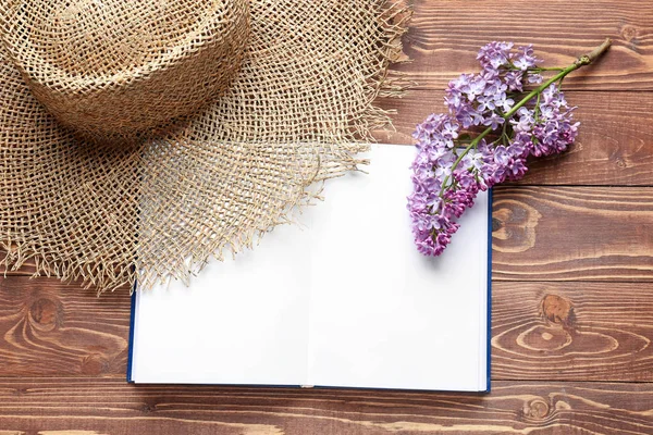 Open notebook, beautiful lilac flowers and hat on wooden background — Stock Photo, Image