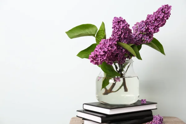 Beautiful lilac flowers in vase with books on table against light background