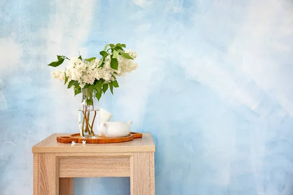 Beautiful lilac flowers in vase with teapot on table against color background — Stock Photo, Image