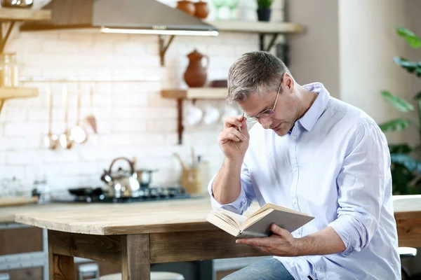 Schöner reifer Mann liest Buch in Küche zu Hause — Stockfoto